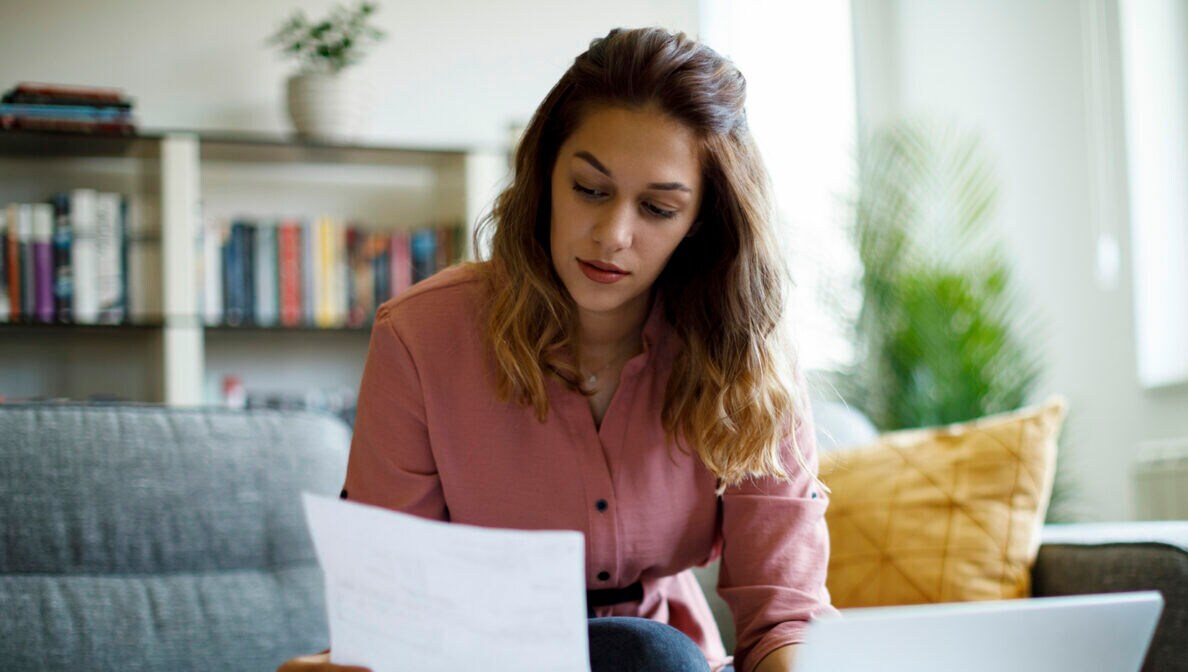Eine Frau sitzt am Tisch vor ihrem Laptop und hält ein Dokument in den Händen