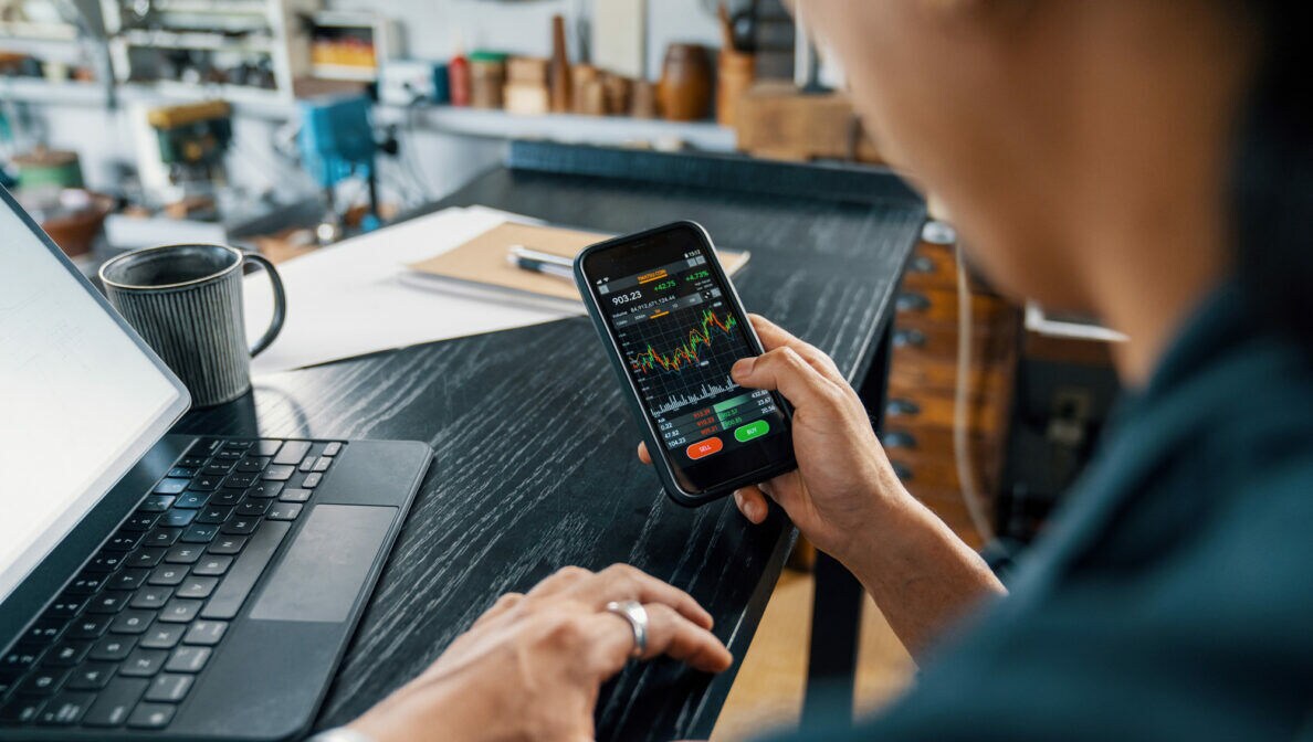 Eine Person mit Smartphone in der Hand, auf dem Aktienkurse zu erkennen sind