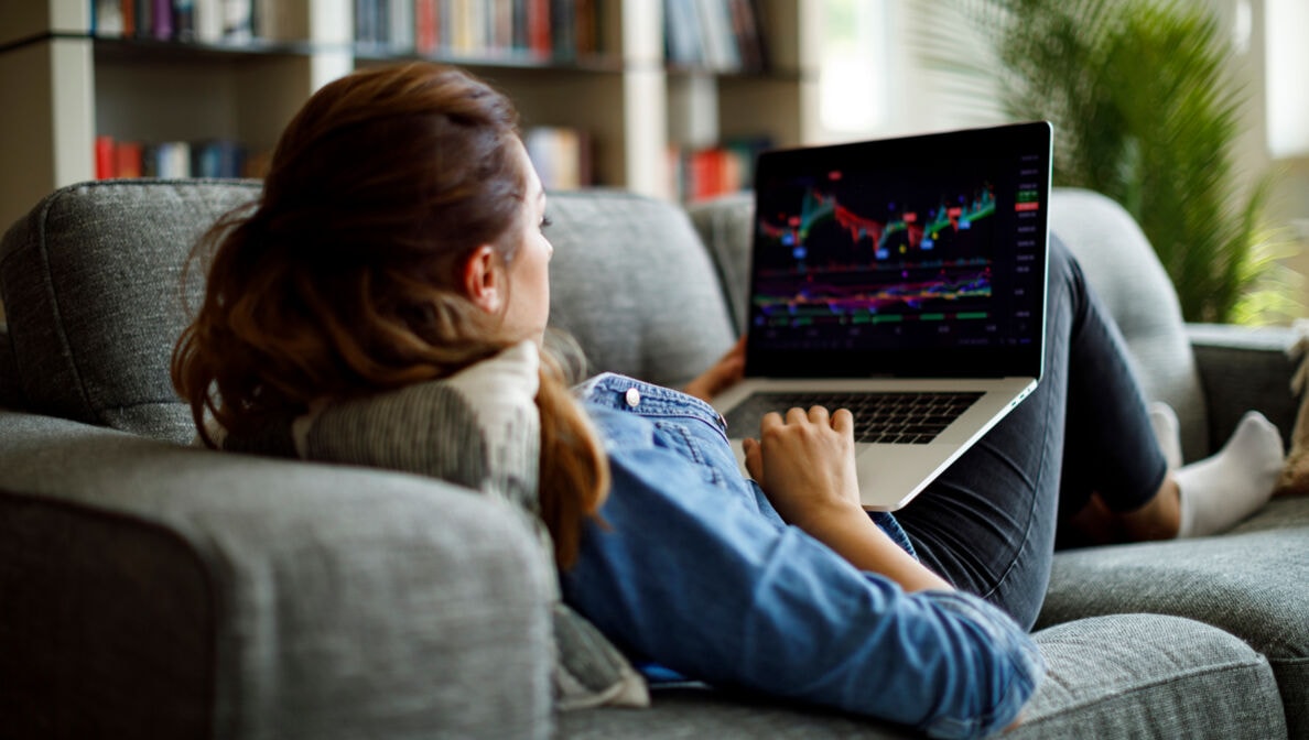 Eine junge Frau liegt mit einem Laptop im Schoß auf einer Couch und schaut auf Aktiencharts.