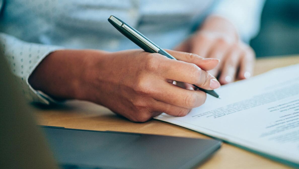 Person im hellen Businesshemd sitzt am Schreibtisch und unterschreibt ein Dokument