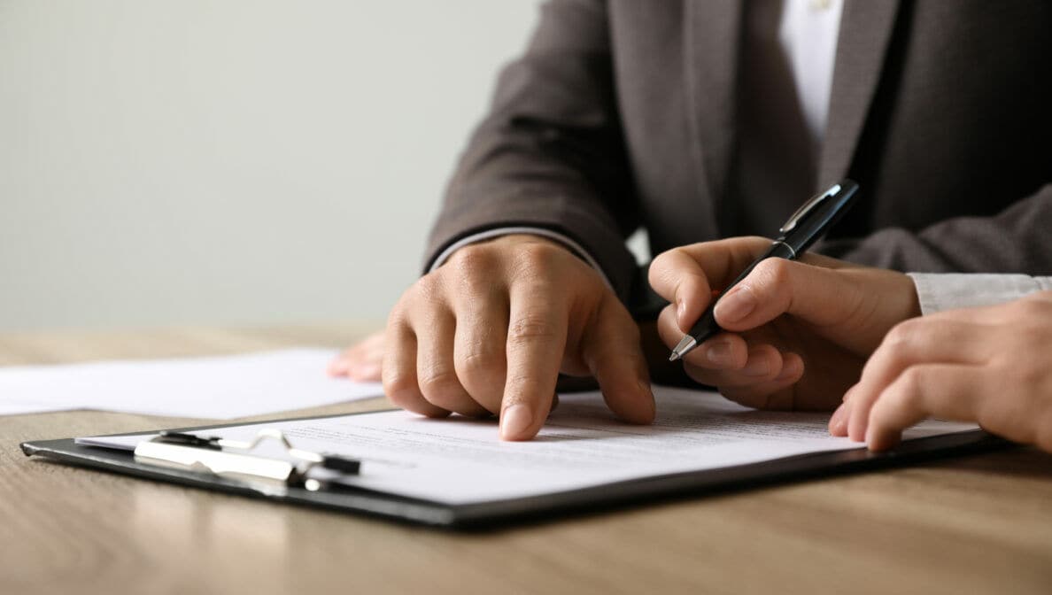 Zwei Personen in Businesskleidung über einem Dokument; die eine Person deutet auf eine Passage, während die andere einen Kugelschreiber hält.