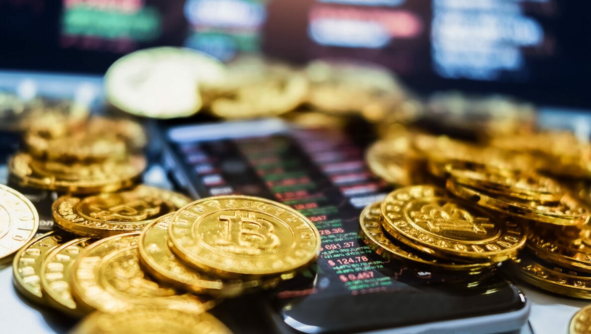 Bitcoin-Münzen liegen auf einem Smartphone, das Börsenkurse zeigt.
