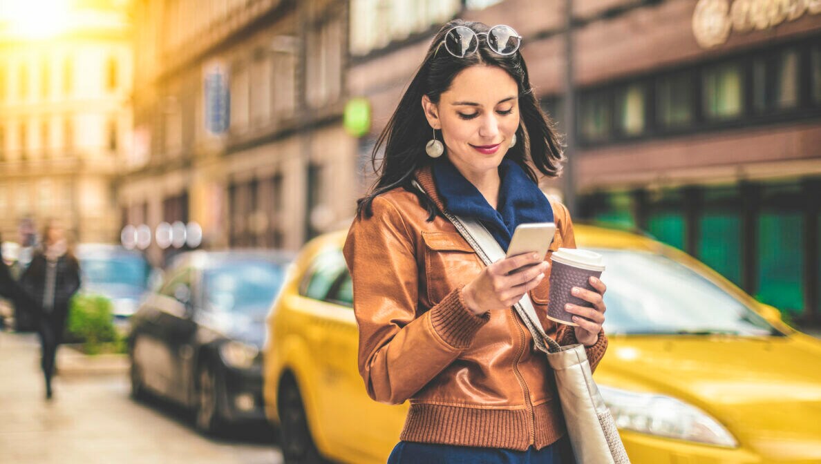 Eine Frau steht vor einem Auto, blickt auf ihr Handy und hält dabei ein Kaffeebecher in der Hand.