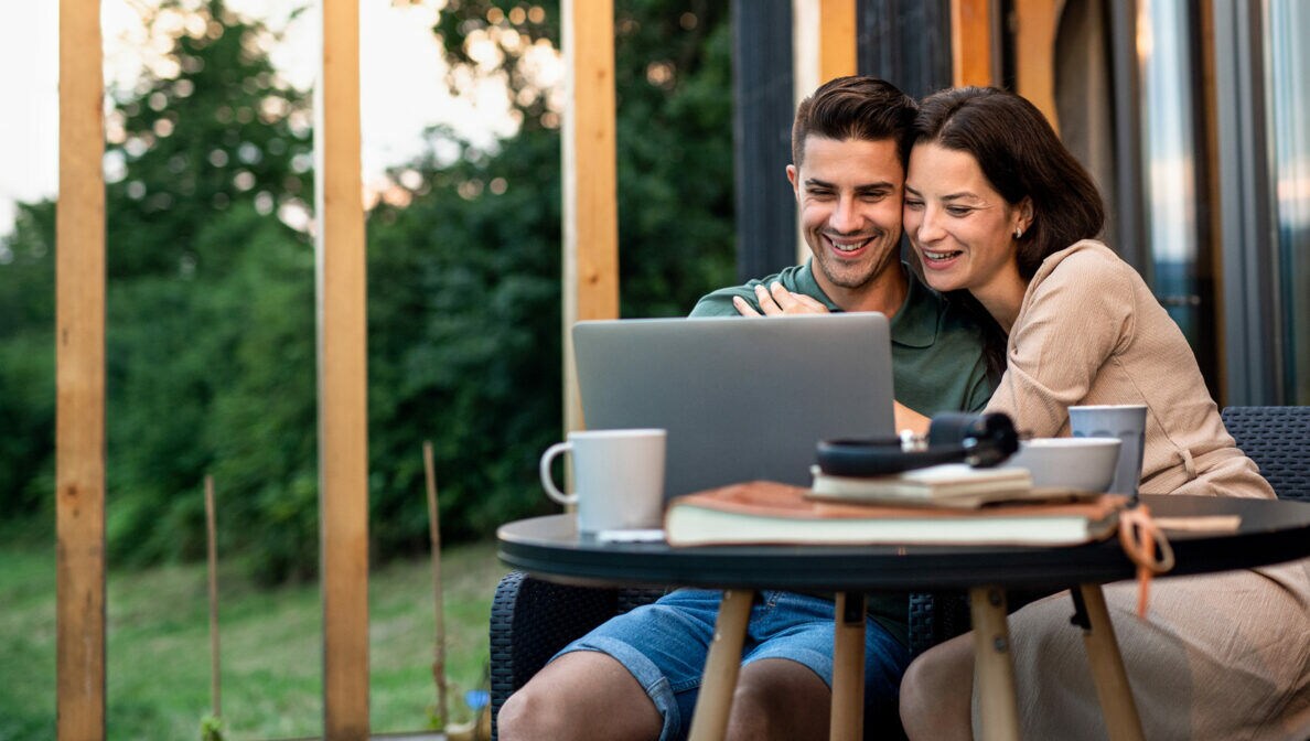 Ein Paar sitzt auf einer Terrasse an einem Tisch und blickt auf einen Laptop.