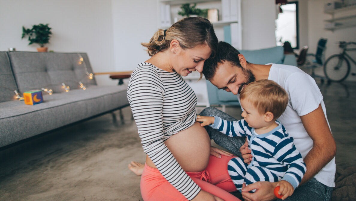 Eine schwangere Frau und ein Mann mit Kleinkind auf dem Schoß, das auf den Babybauch blickt, in einer Wohnung auf dem Teppichboden