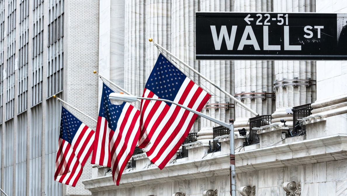 US-amerikanische Flaggen hängen vor einem historischen Gebäude. Im Vordergrund ist das Straßenschild der Wall Street zu sehen. 