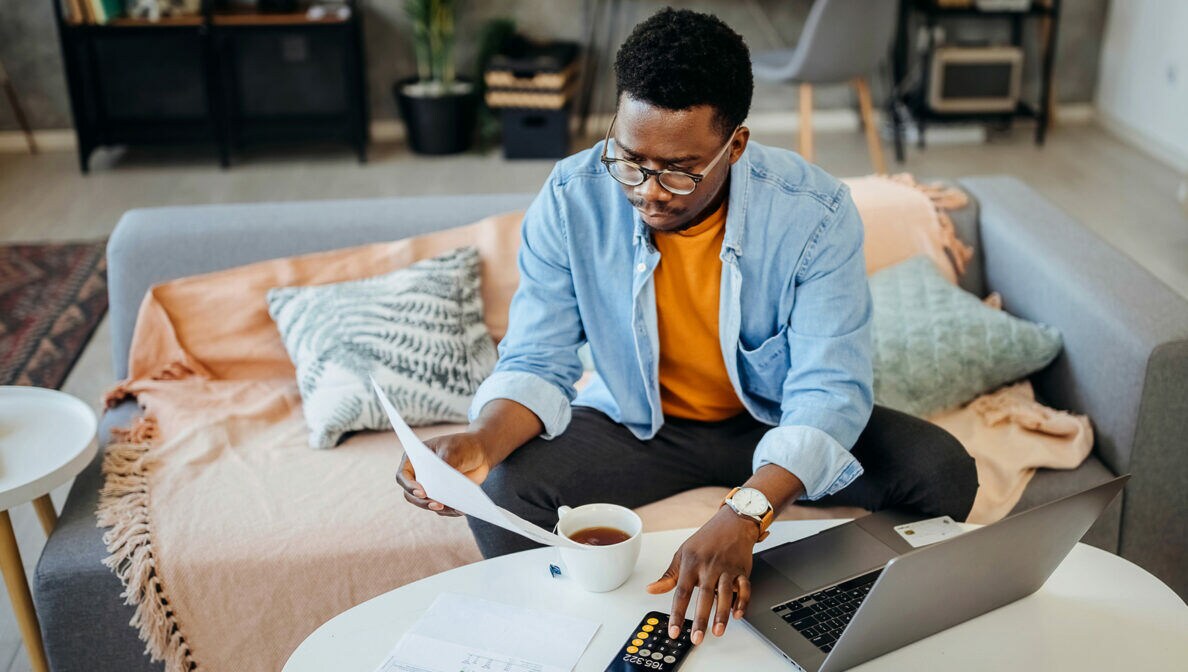 Eine Person sitzt am Couchtisch vor einem Laptop, blickt auf Dokumente und tippt auf seinem Smartphone