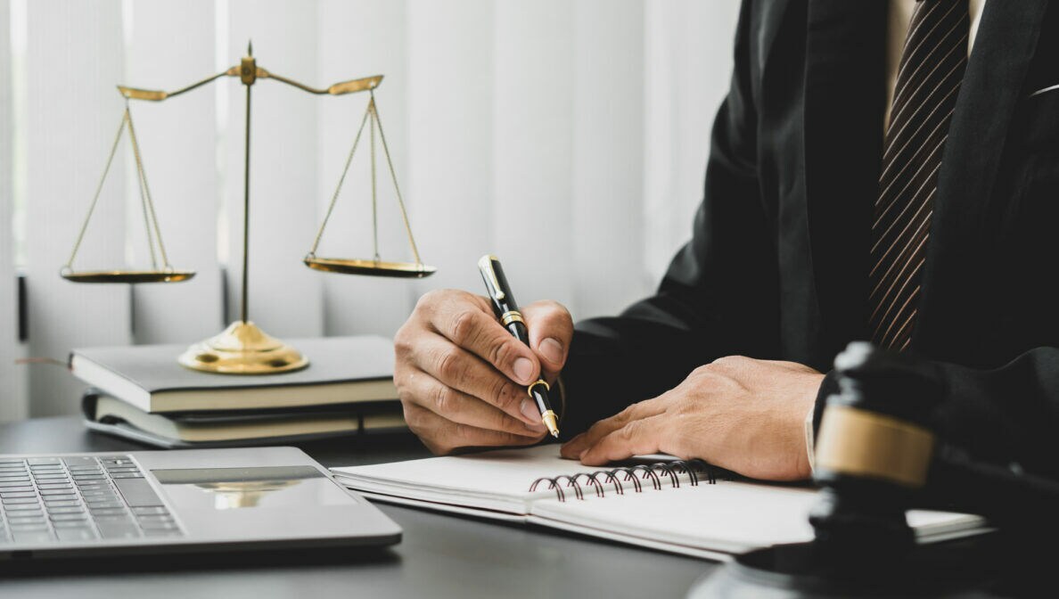 Person im Anzug mit Laptop, Block und Stift bei der Arbeit am Schreibtisch, im Hintergrund steht die Waage der Gerechtigkeit
