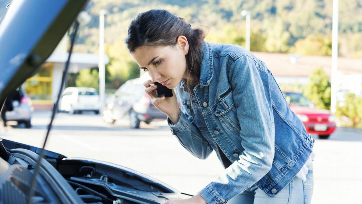 Eine junge Frau steht vor der geöffneten Motorhaube ihres Autos und telefoniert dabei.
