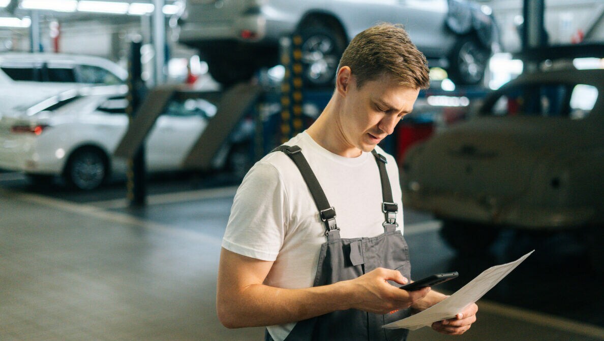 Eine Person in Arbeitskleidung steht in einer Werkstatt und hält ein Smartphone sowie ein Dokument in ihren Händen