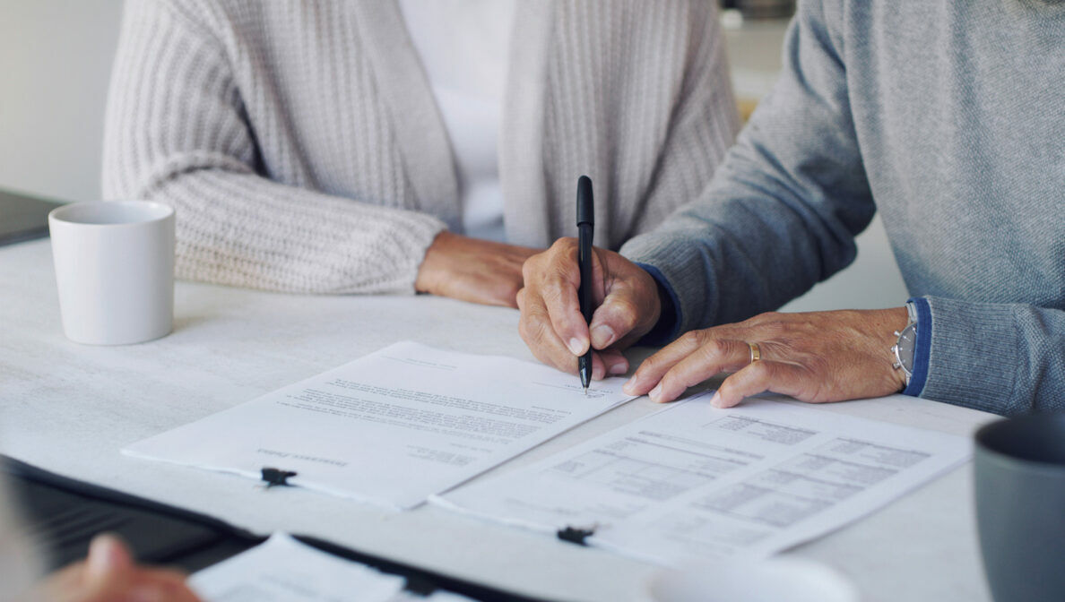 Zwei Personen sitzen an einem Schreibtisch, eine von ihnen unterschreibt Dokumente