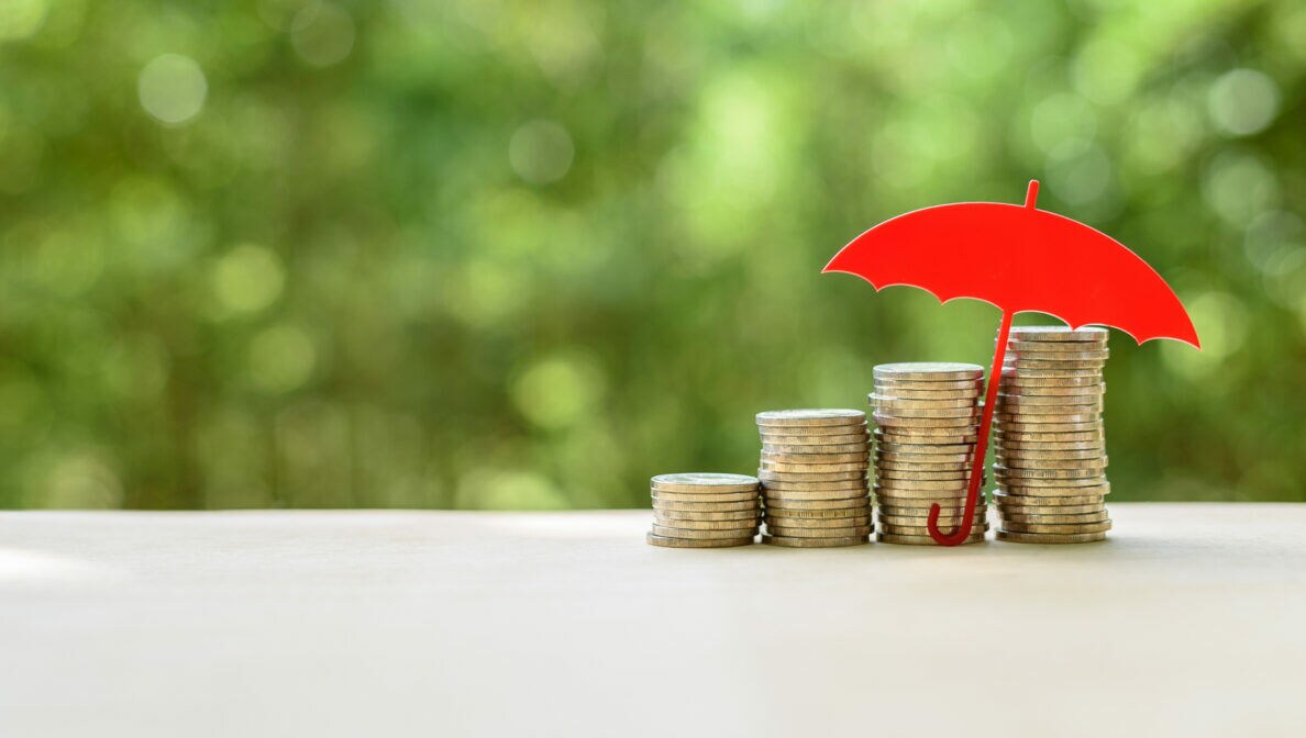 Vier Stapel Münzen, die nach rechts hin höher werden, darüber ein roter 2-D Regenschirm