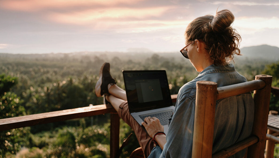 Eine Frau sitzt im Freien auf einer Terrasse, arbeitet an einem Laptop mit Blick ins Grüne.