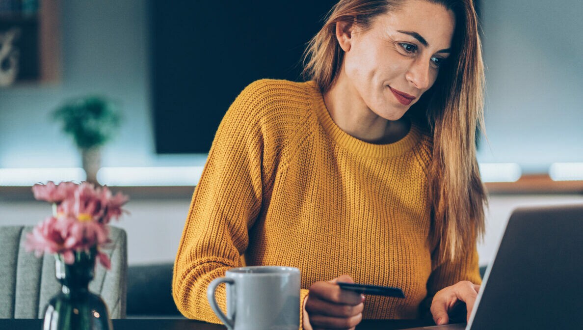 Eine Frau im gelben Pullover sitzt vor einem Laptop und hält eine Bankkarte in der Hand