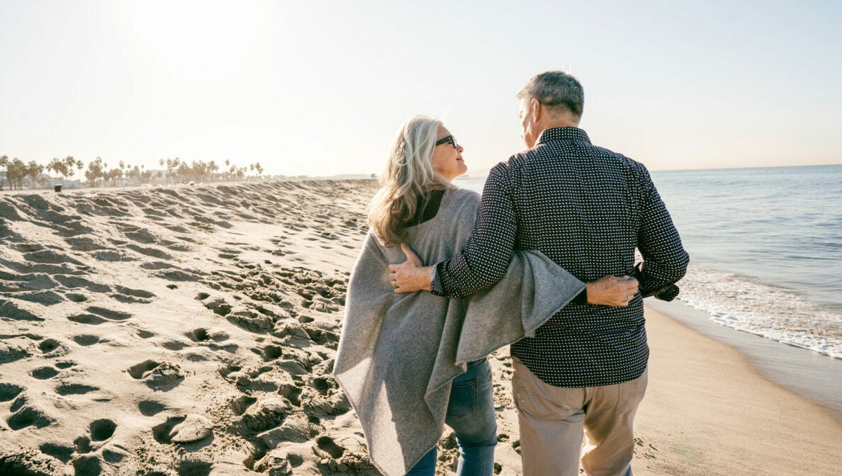 Ein älteres Paar in der Rückansicht spaziert am Strand entlang und hält sich dabei in den Armen