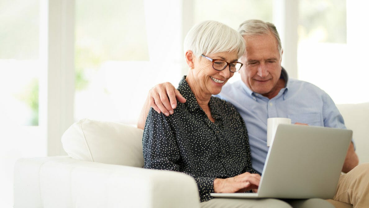 Eine ältere Frau und ein älterer Mann gemeinsam auf dem Sofa und auf ein Laptop blickend, der auf ihren Knien steht