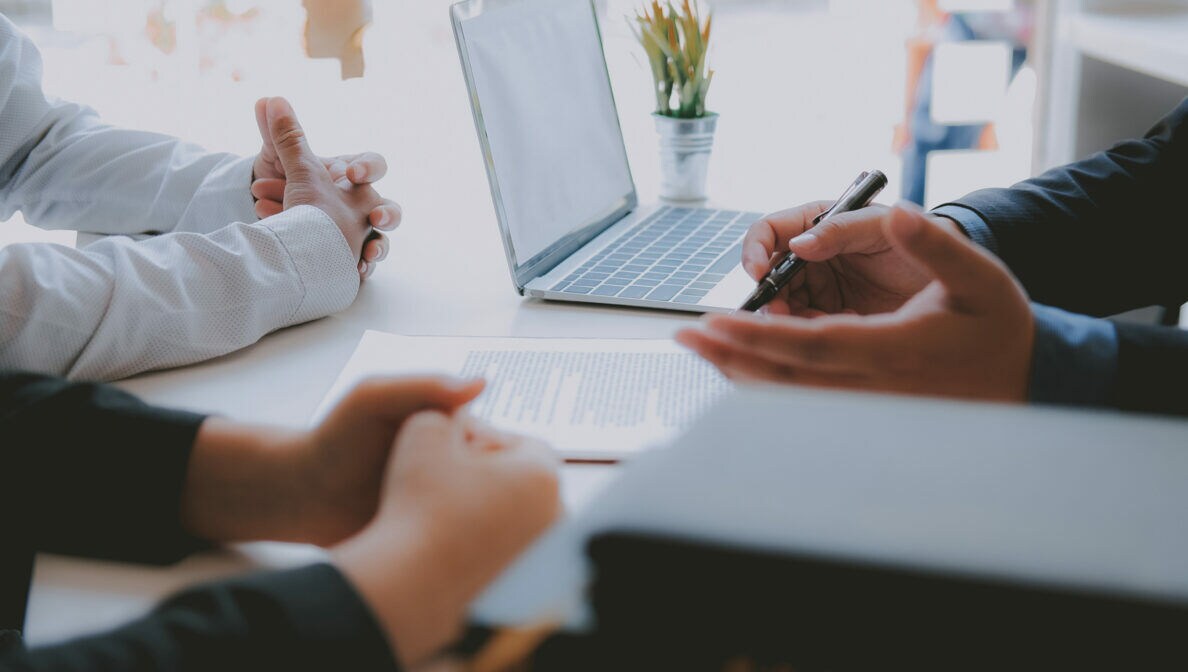 Drei Personen, von denen jeweils nur die Unterarme zu sehen sind, sitzen gemeinsam an einem Tisch, auf dem sich ein Dokument und ein Laptop befinden