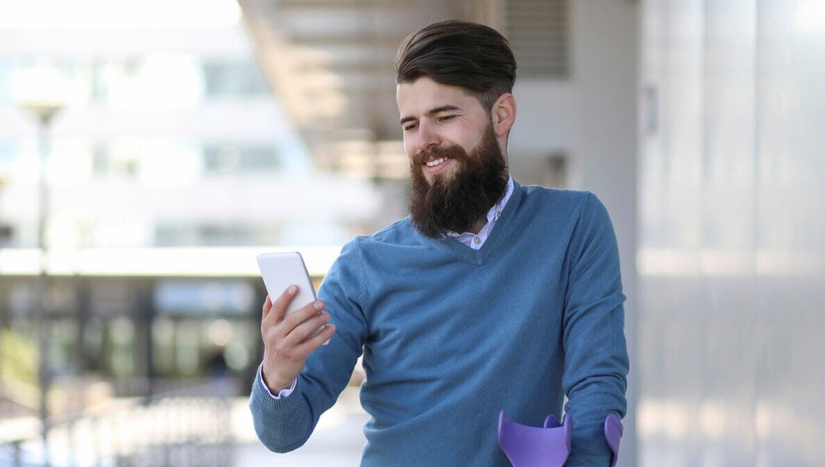 Mann mit Krücken steht vor einem Gebäude und blickt lächelnd auf sein Smartphone.