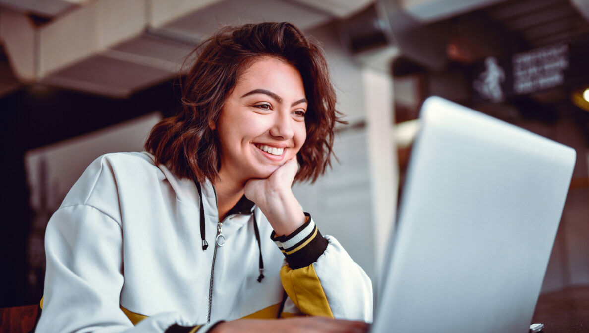 Eine junge Studentin sitzt lächelnd vor einem Laptop in einem Universitätsgebäude.