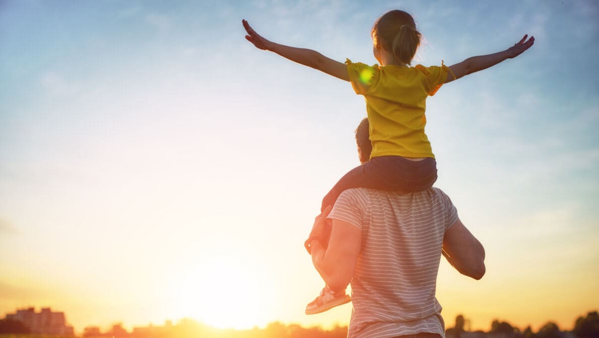 Ein Elternteil trägt ein Kind, das seine Arme ausbreitet, bei Sonnenuntergang auf den Schultern.
