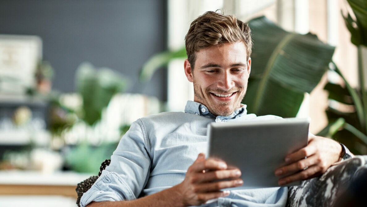 Junger Mann mit blauem Hemd liegt auf einer Couch und schaut auf lächelnd auf ein Tablet.