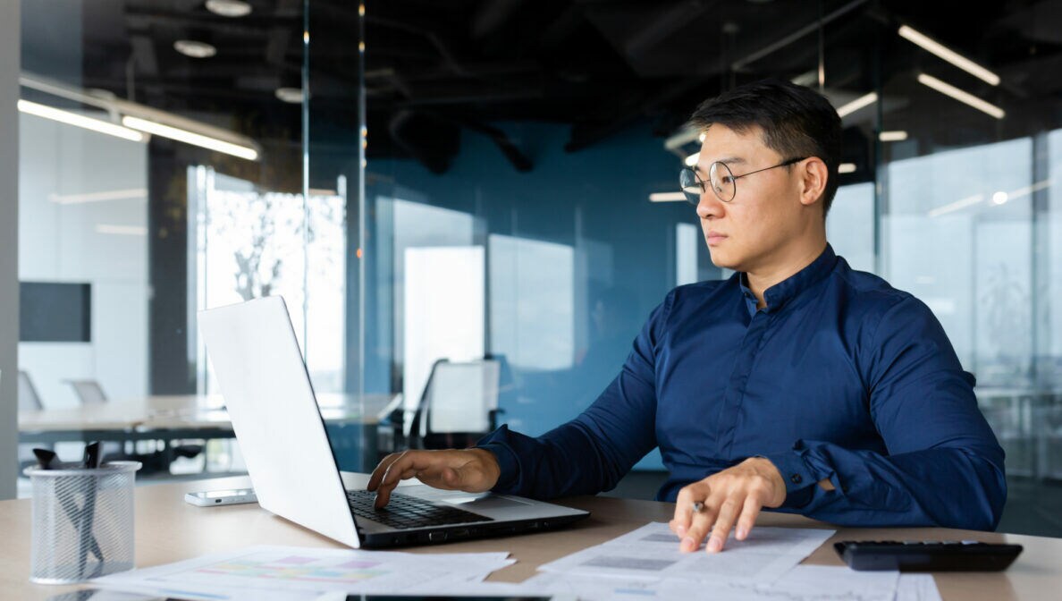 Eine Person sitzt im Büro an einem Schreibtisch mit zahlreichen Dokumenten und blick auf ein geöffnetes Laptop