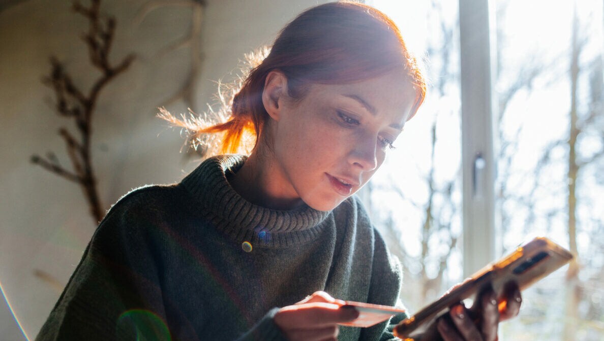 Eine junge Frau mit Zopf und Rollkragenpullover sitzt zu Hause und schaut auf das Smartphone in ihrer linken Hand. In ihrer rechten Hand hält sie eine Bankkarte.