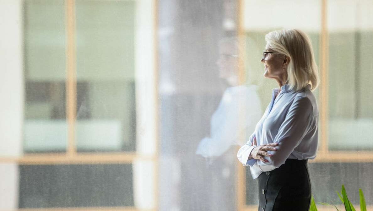 Eine Frau steht mit verschränkten Armen an einer Fensterfront und blickt hinaus