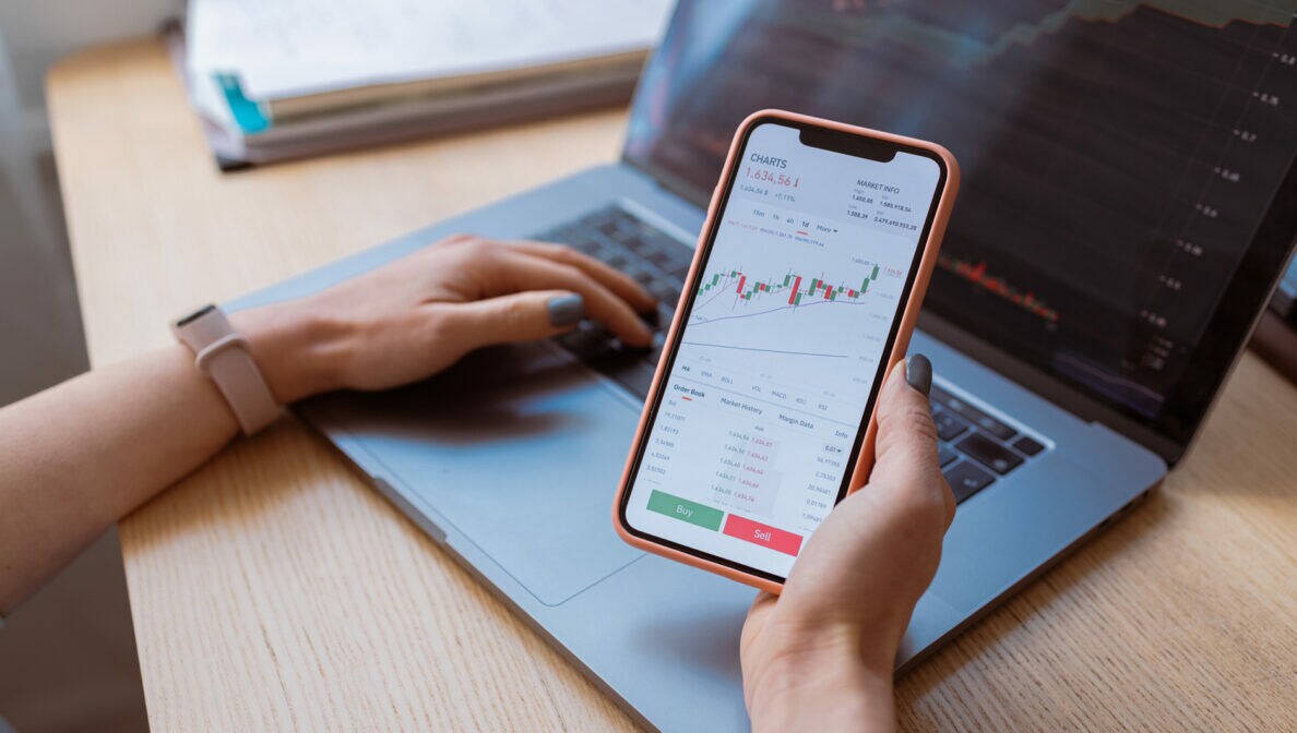 Eine Person sitzt vor einem geöffneten Laptop am Tisch und hält ein Smartphone mit Aktienkursen in der Hand