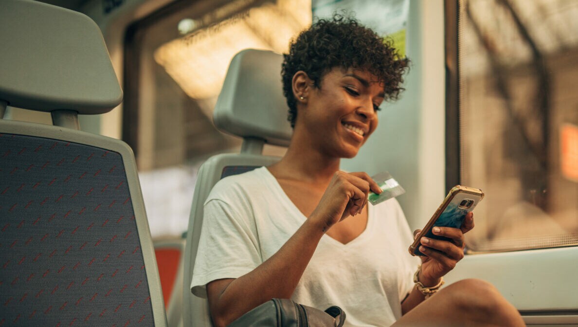 Frau im Zug mit Smartphone in der einen und einer Geldkarte in der anderen Hand