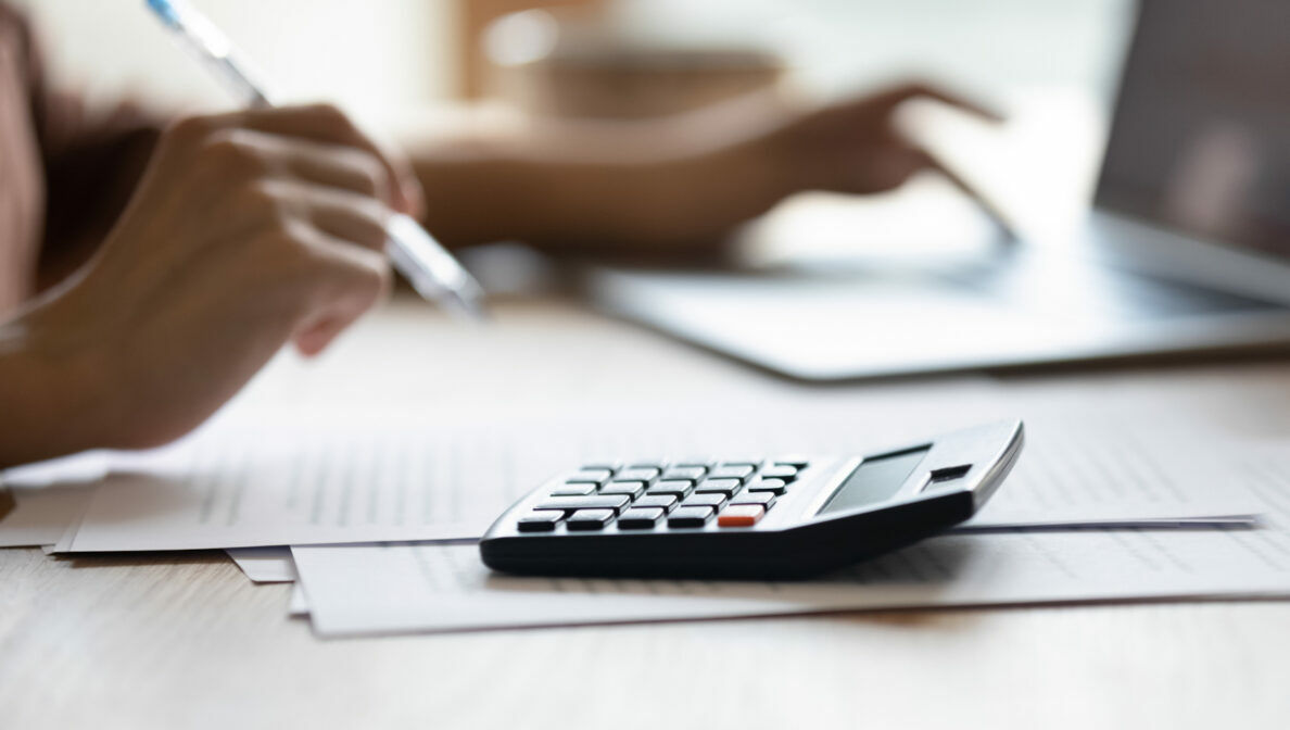 Ein Taschenrechner und Zettel auf einem Schreibtisch neben einem Laptop, auf dem eine Person tippt und in der anderen Hand einen Kugelschreiber hält