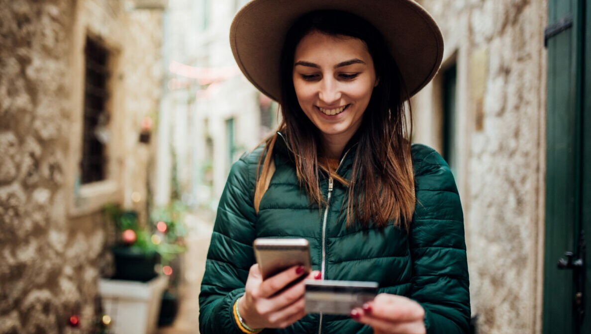 Eine junge Frau mit Hut und grüner Steppjacke beim Online-Shopping mit Smartphone und Bankkarte.