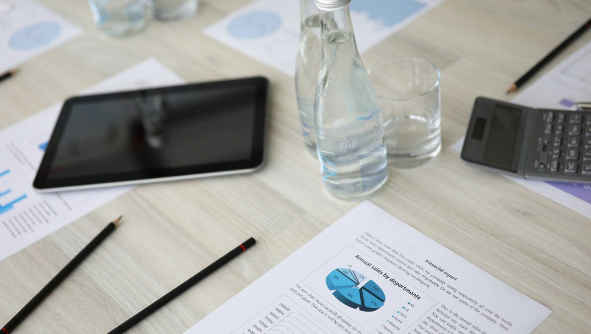 Ein Tablet und Finanzdokumente sowie ein Taschenrechner, Stifte, Wasserflaschen und Gläser auf einem Tisch