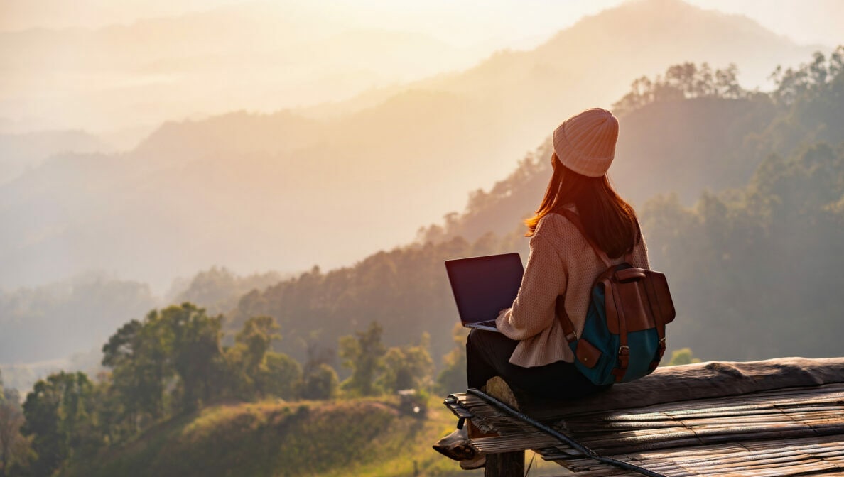 Eine Person mit Laptop sitzt auf einem hölzernen Untergrund und blickt auf ein Naturpanorama mit Bergen und Sonnenaufgang