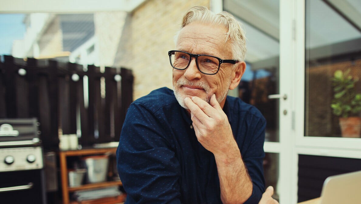 Eine Person sitzt mit Kaffee und geöffnetem Laptop am Tisch auf einer überdachten Terrasse