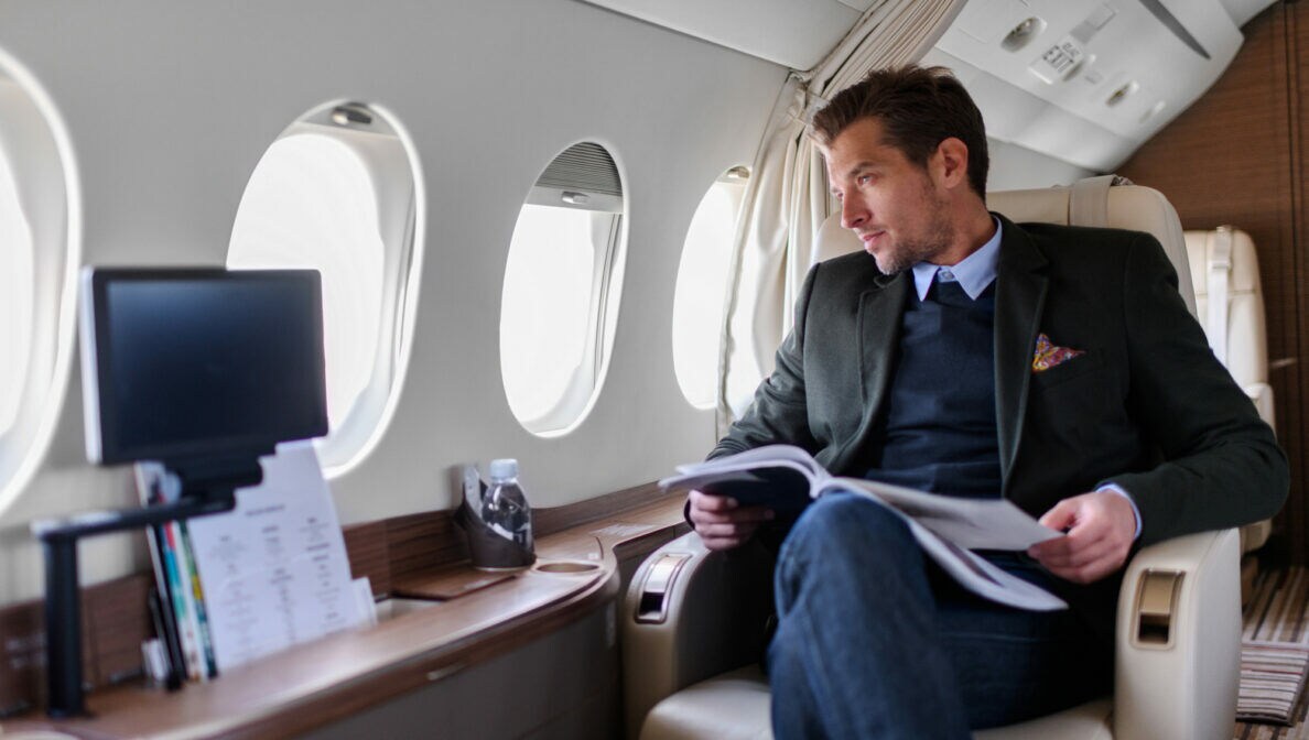 Eine Person sitzt im Privatjet, blickt aus dem Fenster und hat ein Magazin auf dem Schoß liegen