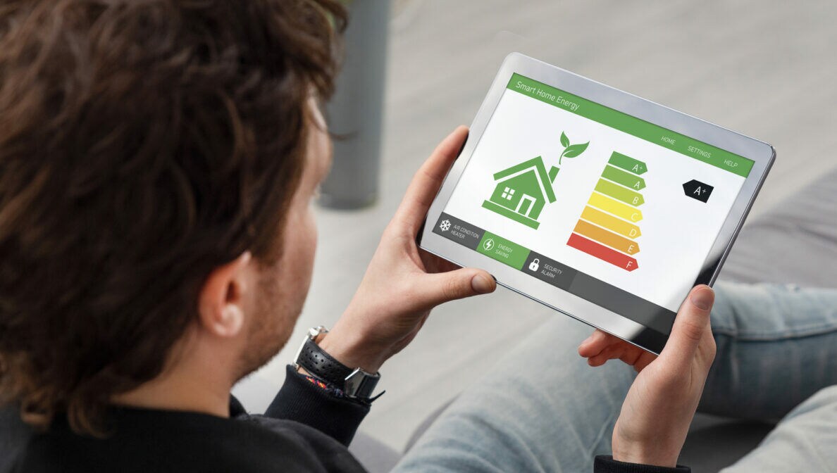 Blick über die Schulter einer Person, die ein Tablet in den Händen hält, auf dem Energieeffizienzklassen und de schematische Darstellung eines Hauses abgebildet sind.