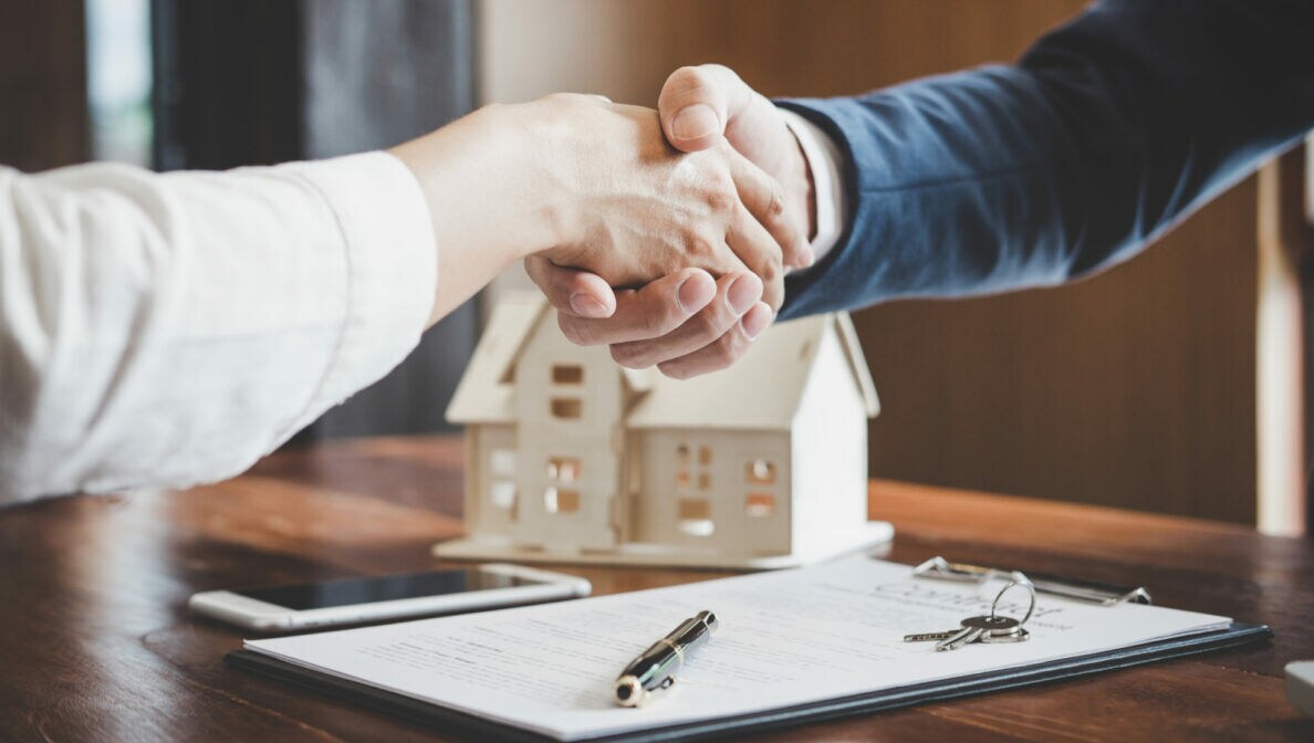 Zwei sich schüttelnde Hände über einem Dokument, im Hintergrund steht ein Modellhaus aus Holz