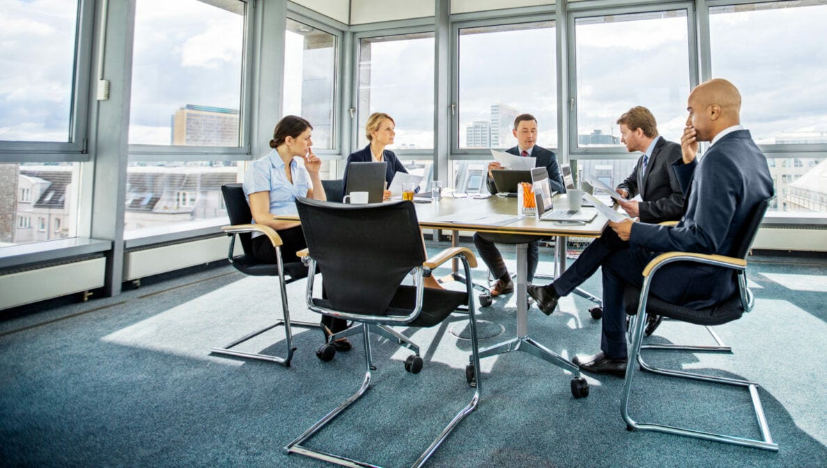 Fünf Personen, drei Männer und zwei Frauen, sitzen rund um einen Tisch in einem hellen Konferenzraum