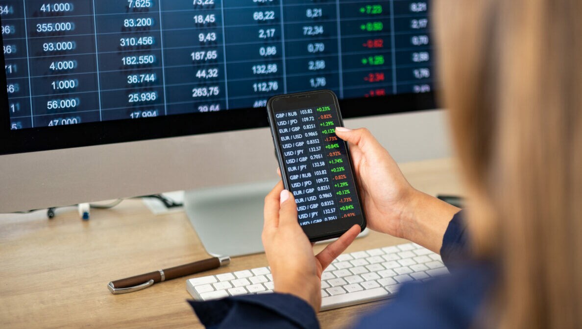 Einer Person wird über die Schulter geblickt, wie sie auf einen Monitor sowie ein Smartphone-Display mit Börsenzahlen schaut