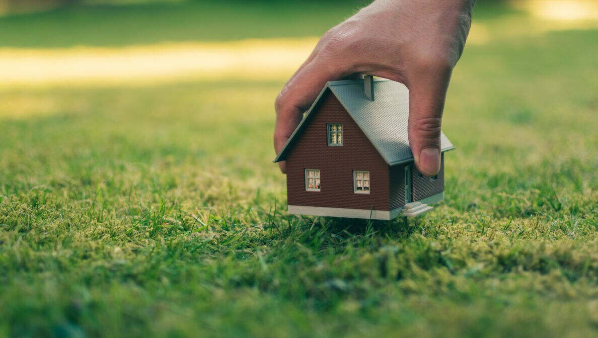 Eine Hand hält ein kleines Modellhaus über eine Rasenfläche
