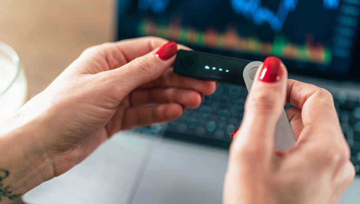 Weibliche Hände halten eine Hardware Wallet vor einem Laptop hoch