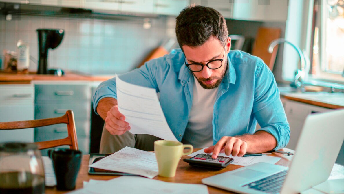 Eine Person sitzt am Küchentisch vor Dokumenten und tippt auf einem Taschenrechner