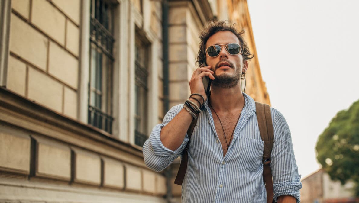 Mann mit Bart, Sonnenbrille und Rucksack auf dem Rücken steht auf einem Bürgersteig und telefoniert