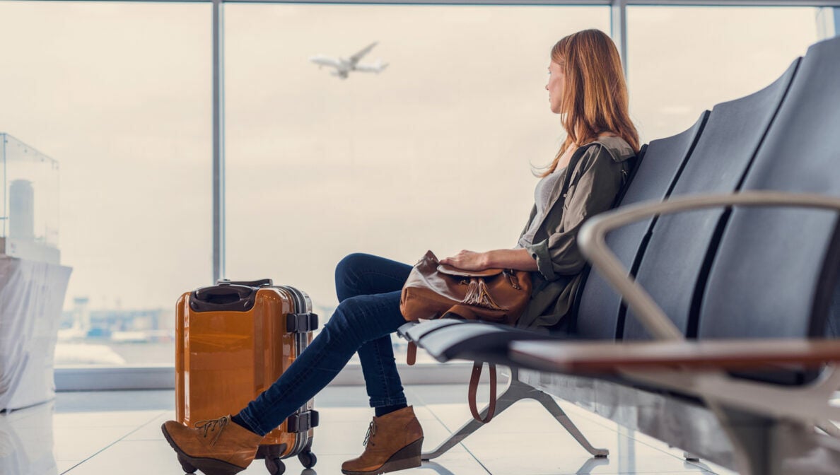 Eine Person sitzt mit einem Koffer im Flughafen und blickt aus dem Fenster, an dem gerade ein Flugzeug vorbeifliegt