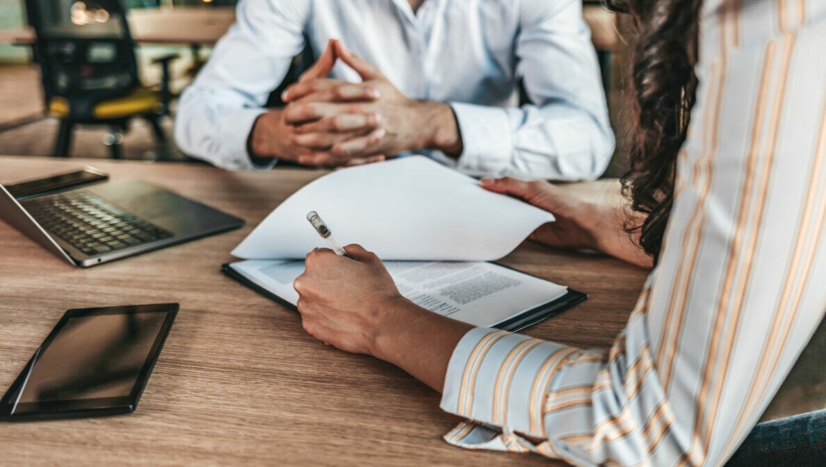 Zwei Personen sitzen an einem Tisch, auf dem ein Laptop und ein Tablet liegen. Eine der Personen hat einen Stift in der Hand und blättert in Unterlagen.