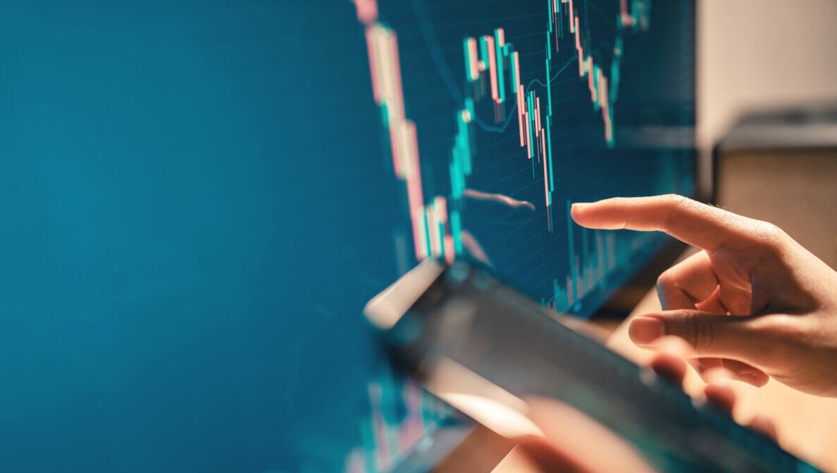 Ein Bildschirm, auf dem Aktienverläufe zu sehen sind. Davor eine Hand, die auf den Kurs zeigt.