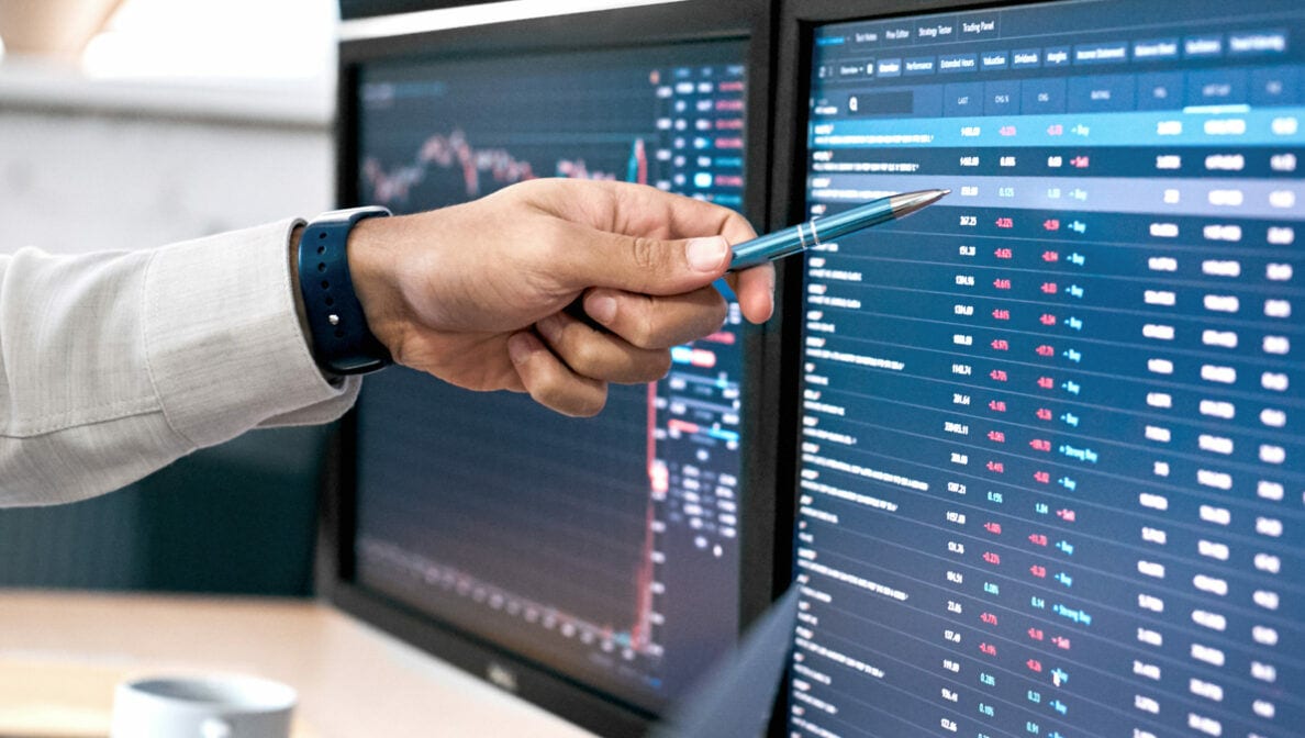 Die Hand einer Person hält einen Kugelschreiber in der Hand und zeigt damit auf einen Bildschirm, auf dem Aktienkurse und -werte zu sehen sind.
