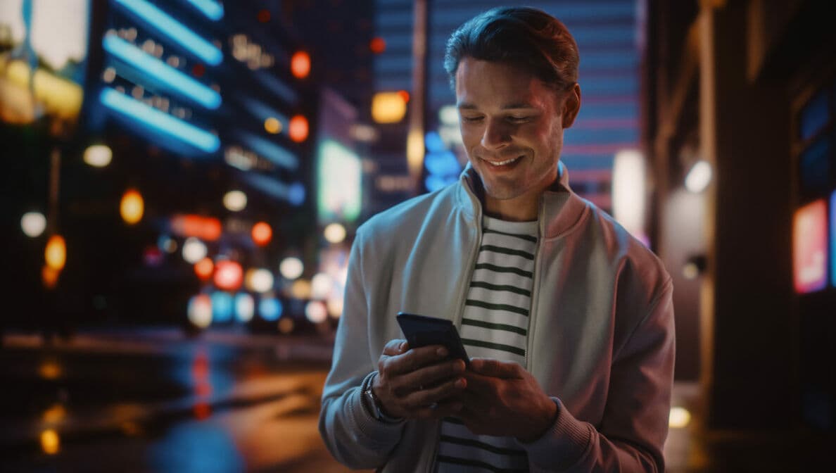 Ein Mann steht auf der Straße einer nächtlich beleuchteten Stadt und schaut lächelnd auf das Smartphone in seiner Hand.