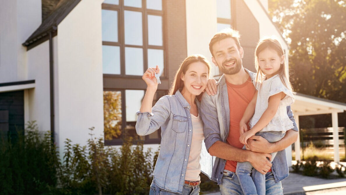 Ein Mann und eine Frau stehen vor einem Haus. Der Mann hält ein Mädchen auf dem Arm, die Frau hält einen Schlüssel in die Höhe.
